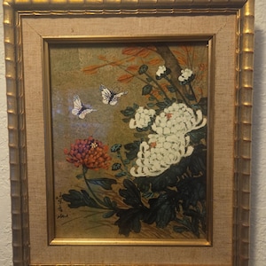 Puede incluir: Pintura floral enmarcada con crisantemos blancos y naranjas, mariposas y follaje. La obra de arte está ambientada sobre un fondo dorado y enmarcada en un marco dorado texturizado. La pintura tiene un estilo tradicional de inspiración asiática.