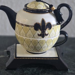 Puede incluir: Juego de té decorativo que incluye una tetera y una taza. La tetera es blanca con un patrón de diamantes amarillos y un diseño de flor de lis negro. El asa y el pico son negros. El juego incluye un soporte negro con la parte superior blanca.