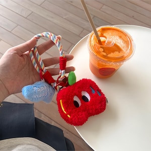 May include: A plush red apple-shaped keychain with a green stem and cartoon eyes. It hangs from a colorful striped lanyard with a blue cloud charm that says "TODAY". A clear plastic cup of orange juice sits on a white table.