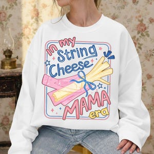 Könnte beinhalten: Weißes Sweatshirt mit einem grafischen Design mit Käsesnacks und dem Text "In my String Cheese Mama Era" in Rosa und Blau. Das Design beinhaltet Sterne und eine Schleife.