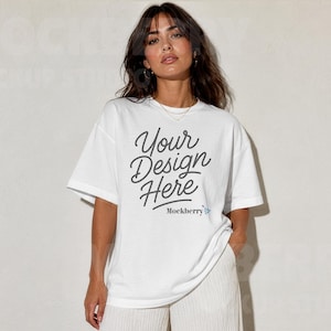 May include: A white short-sleeved t-shirt featuring the dark gray text "Your Design Here" printed in a cursive script on the chest, with the smaller text "Mockberry" and a small blue flower icon positioned directly below the main phrase.