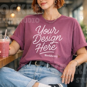 May include: A maroon t-shirt with rolled-up sleeves and the text "Your Design Here" in white script. The person is wearing blue jeans and holding a clear plastic cup with a pink drink and a straw.