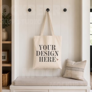 Puede incluir: Un bolso de tela color crema colgado de un gancho de pared negro. El bolso presenta el texto gris oscuro "YOUR DESIGN HERE" impreso en la parte frontal, acompañado de un pequeño gráfico de un triángulo de billar junto a la palabra "HERE".