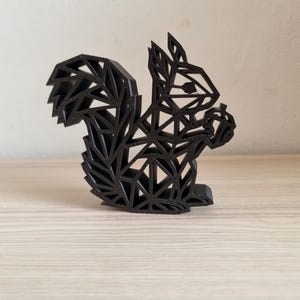 May include: A black, three-dimensional squirrel figurine constructed from a series of interconnected geometric lines and triangular shapes. The squirrel is depicted in a seated position, holding a small nut-shaped object between its front paws.