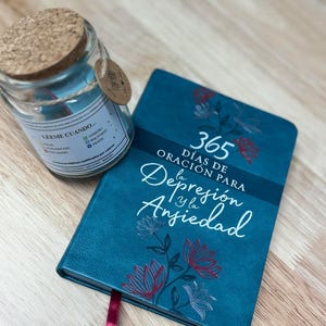 May include: A teal journal with the text "365 Days of Prayer for Depression and Anxiety" and floral accents. A glass jar with a cork lid and a tag is next to the journal. The jar has a label with Spanish text.