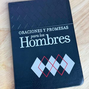 May include: A black notebook with the text "ORACIONES Y PROMESAS para los Hombres" in white. The cover features a diamond pattern and a design of overlapping diamonds in red and white.