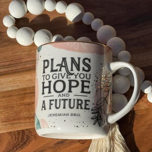 May include: White ceramic mug with the text "PLANS TO GIVE YOU HOPE AND A FUTURE" and the reference "JEREMIAH 29:11". The mug has a gold rim and floral accents. A white tassel and beaded garland are also visible.