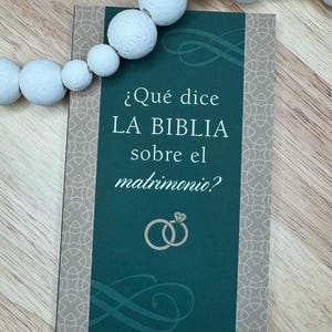 May include: A teal card with the text "¿Qué dice LA BIBLIA sobre el matrimonio?" and two intertwined rings. The card has a beige patterned border and is next to a string of white wooden beads.