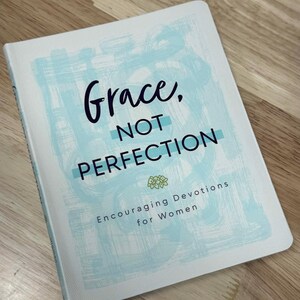 May include: A hardcover book with the title "Grace, Not Perfection" in black script and block letters. The cover features a light blue watercolor design and the text "Encouraging Devotions for Women".