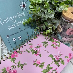 May include: A light blue book titled Confía en Dios con cada aliento, a pink spiral-bound notebook with floral patterns and the word NOTES, and a clear glass tumbler with a bamboo lid and straw, featuring floral graphics and Spanish text.