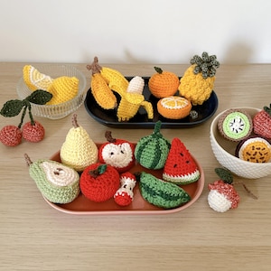 Op de afbeelding: Een verzameling handgemaakte gehaakte vruchten en groenten, waaronder een gele banaan, sinaasappel, ananas, rode kersen, groene peer, rode appel, watermeloenpartje, kiwi en passievrucht, gerangschikt op kleine bordjes en in schaaltjes.