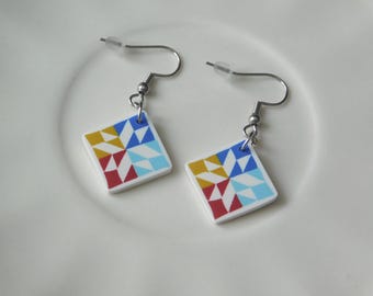 May include: A pair of dangle earrings featuring square white charms with a geometric quilt-inspired pattern in red, yellow, light blue, and dark blue. The charms hang from silver-toned metal fishhook wires with clear plastic earring backs.