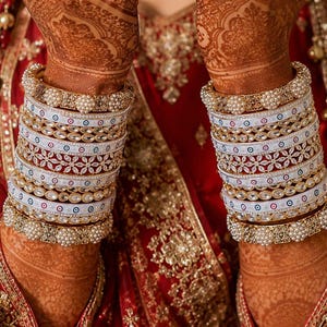 May include: A set of stacked bangles featuring white enamel bands with small circular blue and red accents, interspersed with gold-toned metal bands adorned with clear crystals and pearls, worn over intricate henna patterns on the forearms.