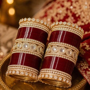 May include: Two wide maroon bangles featuring intricate gold-tone metal detailing. Each bangle is adorned with rows of small clear stones, a central band of circular stone-encrusted medallions, and decorative scalloped edges at the top and bottom.