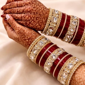 May include: Close-up of ornate red and gold bangles with clear gemstones. The bangles are stacked on a wrist with intricate henna designs. The jewelry is set against a soft, cream-colored background.