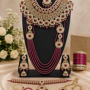Puede incluir: Un conjunto de joyería en tono dorado con piedras de color rojo intenso y perlas blancas. Incluye un collar de varias capas, un maang tikka, pendientes jhumka, brazaletes rojos, una gargantilla, un anillo nasal y dos pulseras de mano.