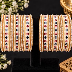 May include: A set of two stacks of bangles featuring alternating bands of gold-tone metal, white enamel with red and blue circular eye motifs, and rows of small white faux pearls and clear rhinestones, arranged in a repeating decorative pattern.