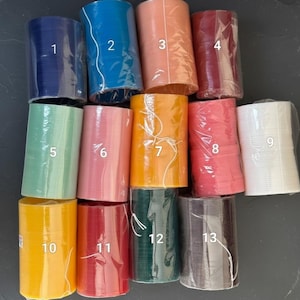 May include: A collection of thirteen rolls of decorative ribbon in various colors, including navy blue, teal, peach, burgundy, mint green, pink, orange, coral, white, yellow, red, dark green, and charcoal gray. Each roll is wrapped in clear plastic.