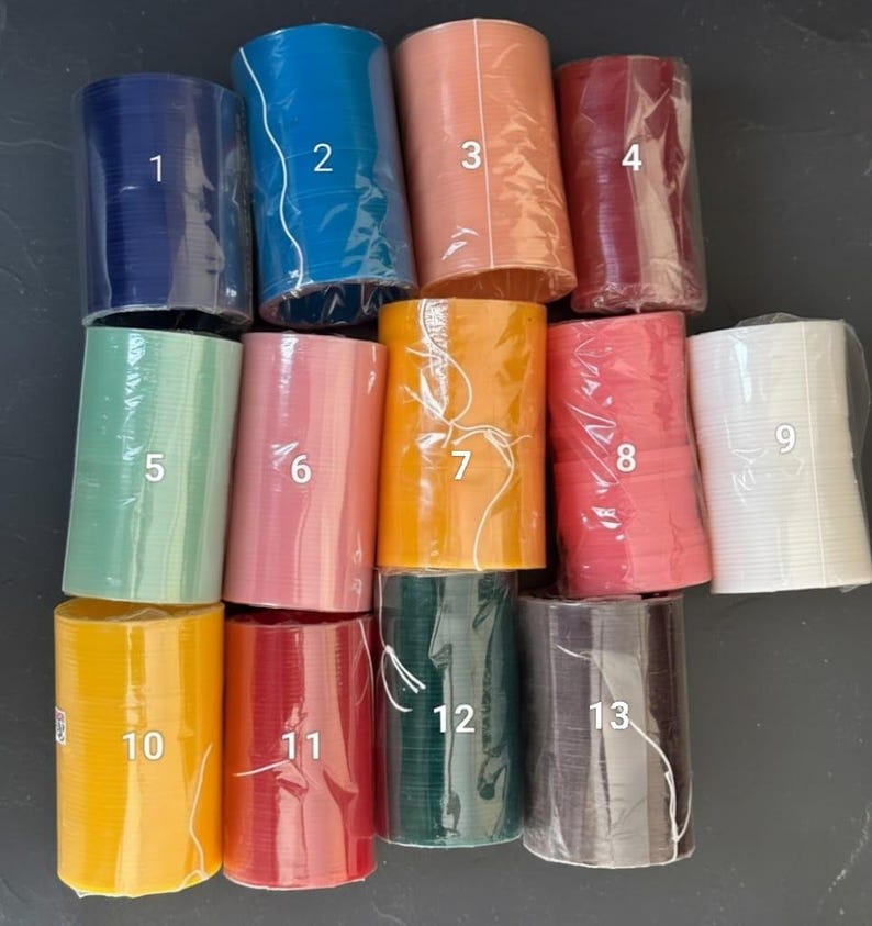 K&ouml;nnte beinhalten: Eine Sammlung von dreizehn Rollen dekorativen Bandes in verschiedenen Farben, darunter Marineblau, T&uuml;rkis, Pfirsich, Burgunderrot, Mintgr&uuml;n, Rosa, Orange, Koralle, Wei&szlig;, Gelb, Rot, Dunkelgr&uuml;n und Anthrazitgrau. Jede Rolle ist in klarem Kunststoff verpackt.