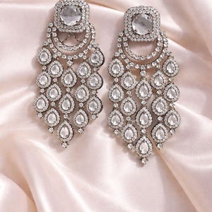 Może przedstawiać: Para srebrnych kolczyków typu żyrandol. Każdy kolczyk ma kwadratową górę z dużym, przezroczystym kamieniem otoczonym mniejszymi kamieniami. Poniżej zwisają liczne kamienie w kształcie łezki, tworząc efekt kaskady.