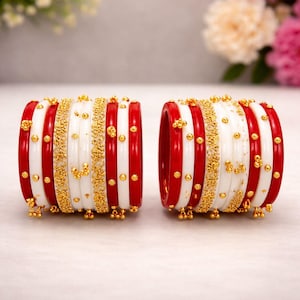 Può includere: Due set di braccialetti rossi, bianchi e dorati. I braccialetti presentano strisce rosse e bianche alternate, con dettagli dorati e piccole perle dorate. Lo sfondo è bianco tenue con fiori sfocati.