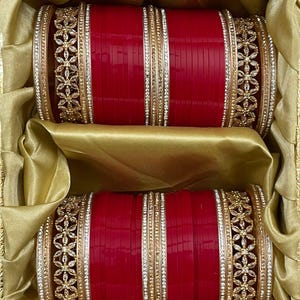 Puede incluir: Dos conjuntos de pulseras rojas con detalles dorados y plateados, presentados en una caja de satén dorado. Las pulseras presentan diseños florales y de pedrería, dando un aspecto lujoso. La caja realza la presentación de las joyas.