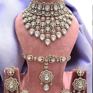 May include: A gold-toned jewelry set featuring a wide, multi-layered choker necklace with clear, oval-shaped stones, matching drop earrings, a central pendant piece with a chain, and a thin circular nose ring, all displaying intricate metalwork.