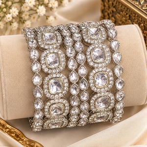 May include: A set of silver-toned bangles featuring large, square-cut clear stones surrounded by smaller, round, pavé-set clear stones. The design includes alternating bands with teardrop-shaped stones, creating a layered, metallic, and sparkling appearance.