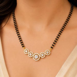 May include: A gold necklace featuring a double strand of small black beads and a central pendant composed of five circular gold frames, each set with clear white stones, with the largest circular frame positioned in the center.