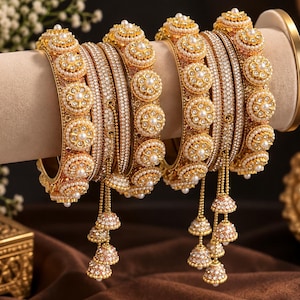 May include: A set of gold-toned bangles featuring intricate floral motifs adorned with small white pearls and clear crystals. The bangles include dangling bell-shaped charms covered in tiny pearls and crystals, creating a layered, ornate appearance.