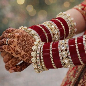 May include: Close-up of hands adorned with intricate henna designs and wearing traditional red and gold bangles. The bangles feature a combination of red, white, and gold elements, including sparkling gemstones and delicate pearl accents.