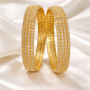 May include: Two gold bangles with a sparkling, diamond-like pattern. The bangles are set against a white, silky background, creating a luxurious and elegant aesthetic. The jewelry is a classic accessory.