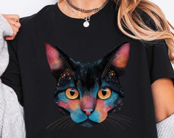 May include: Black t-shirt featuring a colorful cat face design. The cat's face is a mix of blues, pinks, and yellows, resembling a galaxy. The cat has yellow eyes and whiskers. The shirt is a crew neck and short-sleeved.