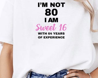 May include: White t-shirt with the text "I'M NOT 80 I AM Sweet 16 WITH 64 YEARS OF EXPERIENCE" in black and pink. The shirt is worn with blue jeans. A woman is wearing the shirt.