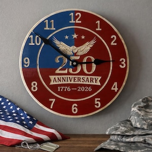 May include: A round wooden clock with a red and blue color scheme. The clock face features the text "250 ANNIVERSARY 1776-2026" and an eagle emblem. The clock is displayed with an American flag and military gear.
