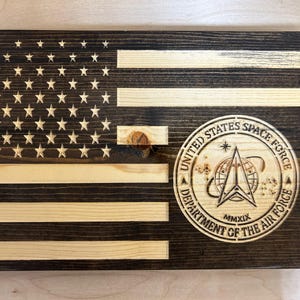 Op de afbeelding: Houten Amerikaanse vlag met het embleem van de United States Space Force. De vlag heeft een donker gebeitste achtergrond met natuurlijke houten strepen en sterren. Het Space Force-embleem is een cirkelvormig ontwerp.