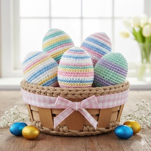Puede incluir: Una cesta de mimbre contiene seis huevos de Pascua de ganchillo a rayas pastel. Los huevos son en tonos de rosa, azul, amarillo y verde. La cesta tiene un forro de cuadros rosa y un lazo a juego.
