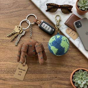 Puede incluir: Una colección de objetos de uso diario sobre una superficie de madera. Incluye un llavero de araña de ganchillo marrón, un llavero de globo de ganchillo azul y verde, llaves, un mando de coche, gafas de sol, una cartera, un teléfono y una etiqueta que dice "Handmade with Love".