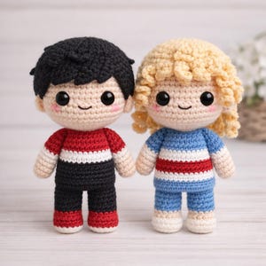 May include: Two handmade crochet dolls. One doll has black hair, a red, white, and black striped shirt, and black pants. The other doll has blonde curly hair, a blue, white, and red striped shirt, and blue shorts. Both dolls have rosy cheeks and smiling faces.