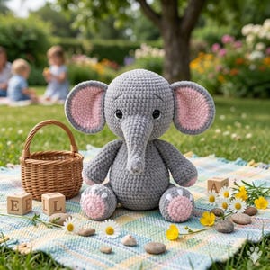 May include: A gray crocheted elephant plush toy with pink inner ears and foot pads sits on a patterned blanket. A small wicker basket and wooden blocks spelling "EEL" are nearby, with daisies and small stones scattered around. The scene is set outdoors.