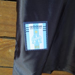 May include: Close-up of a navy blue athletic jersey with a textured pattern. A rectangular patch on the jersey features the Adidas logo, text, and a crest in light blue, white, and gold.