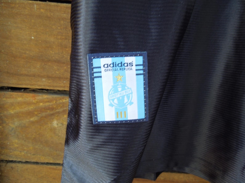 May include: Close-up of a navy blue athletic jersey with a textured pattern. A rectangular patch on the jersey features the Adidas logo, text, and a crest in light blue, white, and gold.