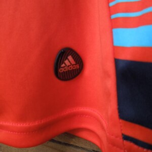 May include: Close-up of a red athletic shirt with the Adidas logo on the sleeve. The shirt has a striped design in red, blue, and navy blue. The Adidas logo is black with the Adidas text in white.