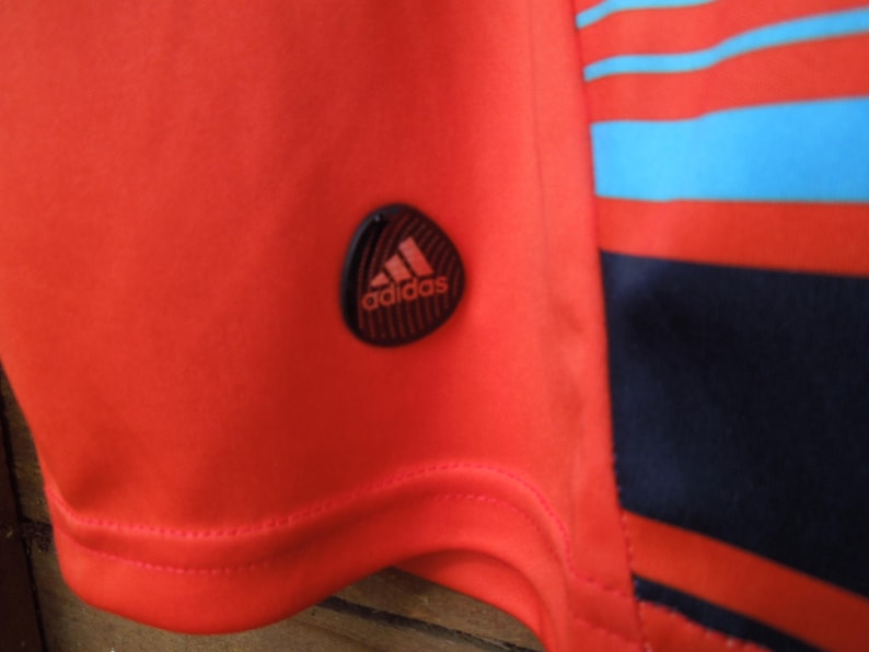 May include: Close-up of a red athletic shirt with the Adidas logo on the sleeve. The shirt has a striped design in red, blue, and navy blue. The Adidas logo is black with the Adidas text in white.