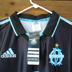 May include: Black and light blue Adidas soccer jersey with the Olympique de Marseille logo and the text "DROIT AU BUT". The jersey has a black collar and light blue stripes. The Adidas logo is visible on the front.