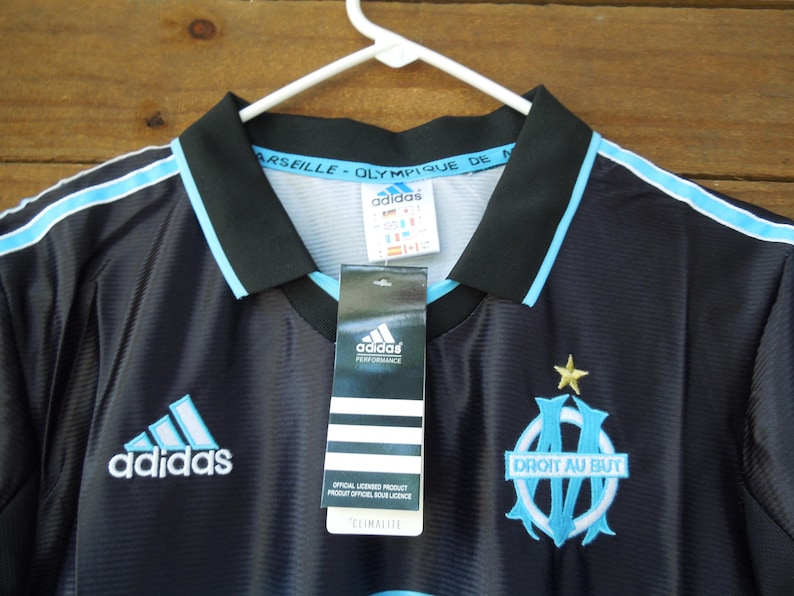 May include: Black and light blue Adidas soccer jersey with the Olympique de Marseille logo and the text "DROIT AU BUT". The jersey has a black collar and light blue stripes. The Adidas logo is visible on the front.