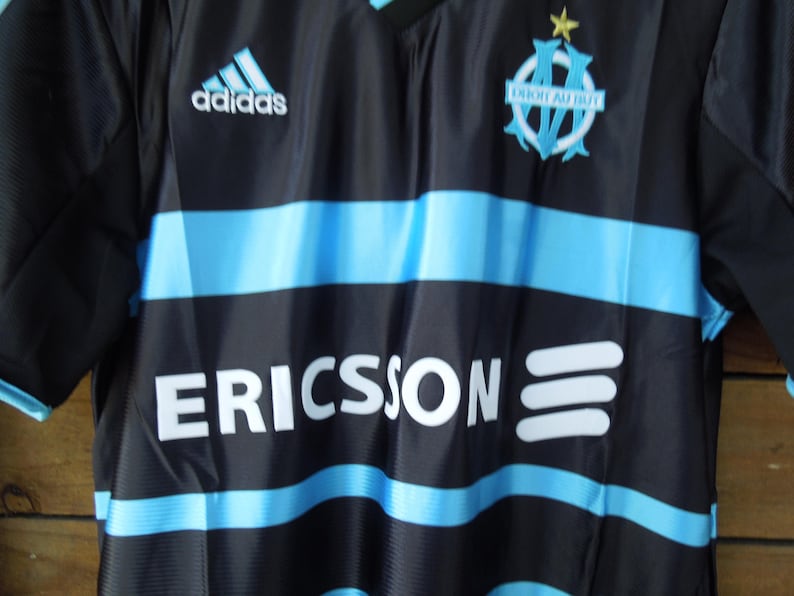 May include: Black and blue soccer jersey with horizontal light blue stripes. Features the Adidas logo, team emblem, and the word "ERICSSON" in white. The jersey has a V-neck design.