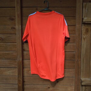 May include: Bright orange athletic shirt with short sleeves, hanging on a black hanger. The shirt has light blue accents on the shoulders and black trim on the sleeves. The shirt is made of a lightweight, breathable material.