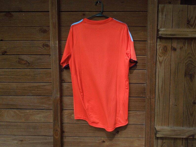 May include: Bright orange athletic shirt with short sleeves, hanging on a black hanger. The shirt has light blue accents on the shoulders and black trim on the sleeves. The shirt is made of a lightweight, breathable material.