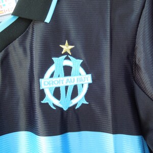 May include: A navy blue and light blue soccer jersey with the emblem of a team. The emblem features a light blue and white design with the words "DROIT AU BUT" and a gold star. The jersey has a V-neck collar.